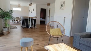 Een ruime woonkamer met houten vloer, een eettafel met stoelen, een ronde rieten fauteuil met kussen, een grijze bank en een grote potplant.