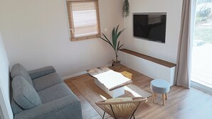 Een moderne woonkamer met een grijze bank, een klein tafeltje, een rieten stoel, een aan de muur bevestigde televisie en een groot raam met jaloezieën.