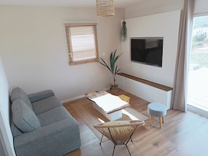 Una sala de estar moderna con un sofá gris, una mesita, una silla de mimbre, un televisor montado en la pared y un ventanal con persianas.