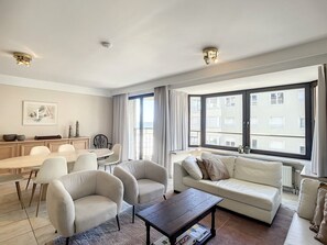 A modern living room with a sofa, armchairs, a coffee table, and a dining area with a table and chairs. - Apartment Near Albert Beach With Terrace (Knokke-Heist)