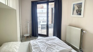 Ein Hotelzimmer mit Bett, Heizkörper, einem Fenster mit Vorhängen, einem Sessel und einem kleinen Tisch.