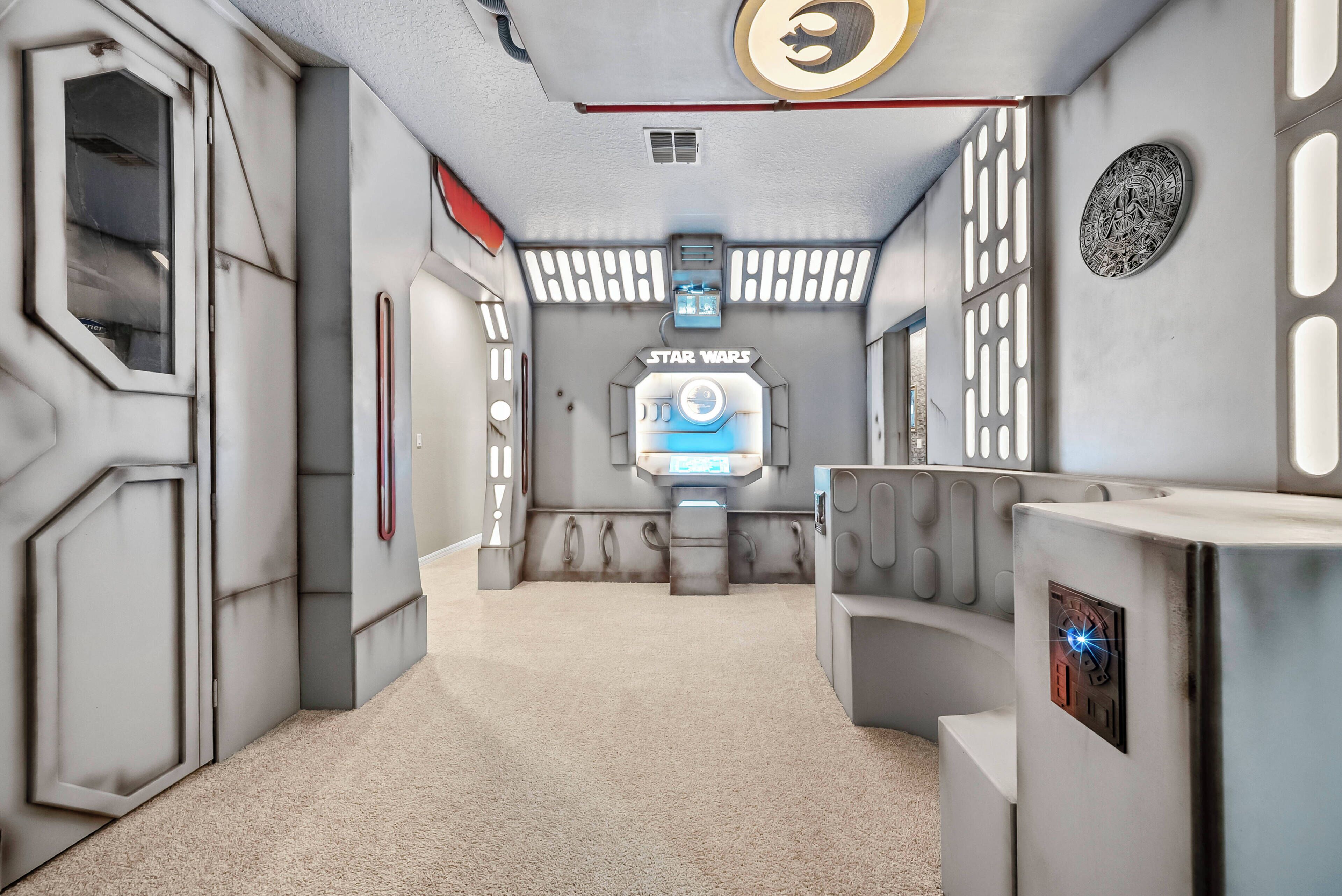 Ett rum med Star Wars-tema, med en central, kapselliknande sittgrupp, en väggmonterad kontrollpanel och ett dekorativt emblem.