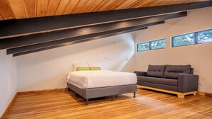 Une chambre à coucher avec un lit, un canapé gris, un plancher en bois, un plafond en pente et deux fenêtres.
