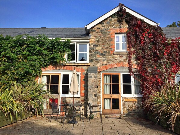 A stone cottage with two windows, a door, and a small patio with a table and chairs.