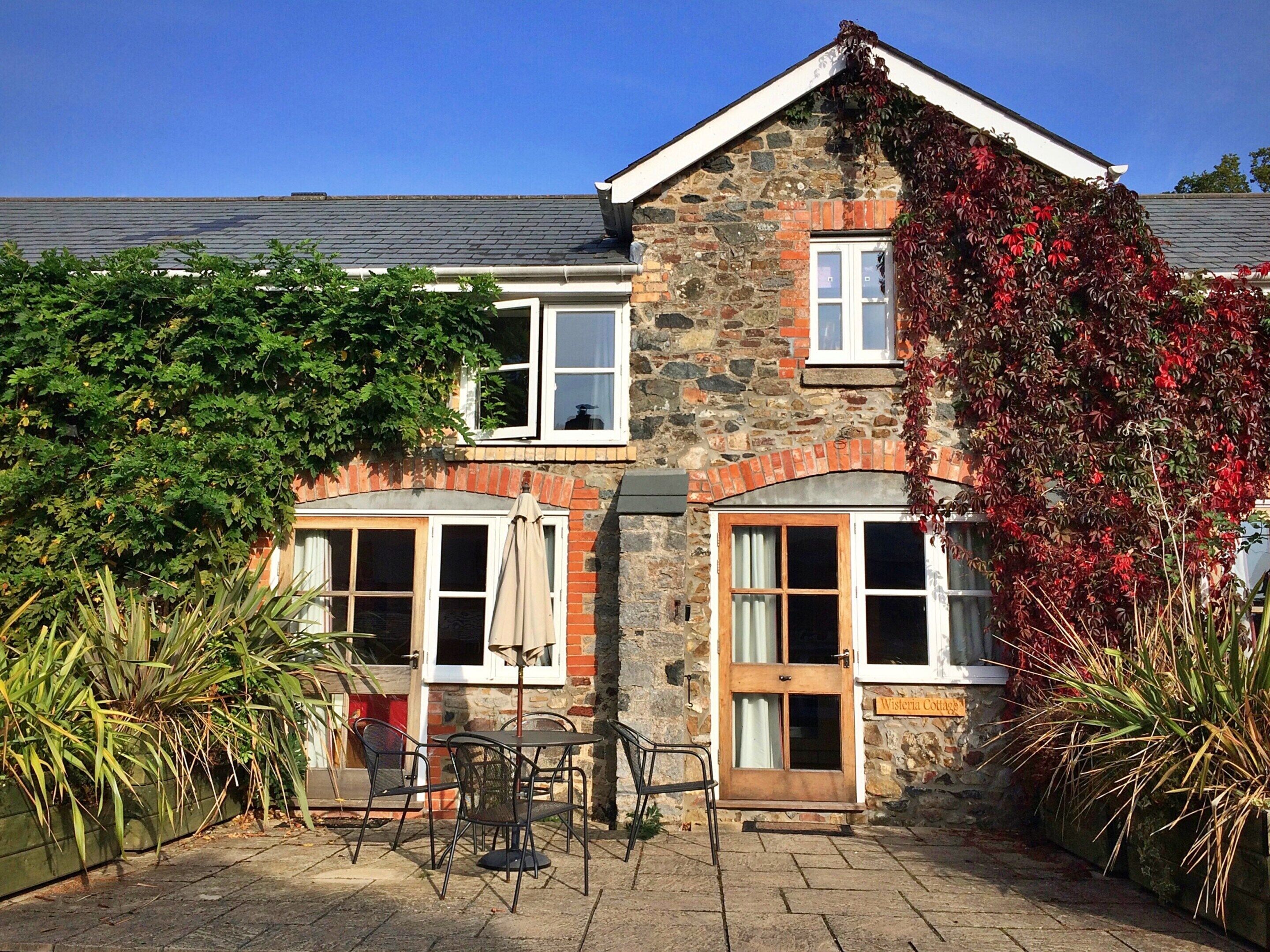 A stone cottage with two windows, a door, and a small patio with outdoor furniture.