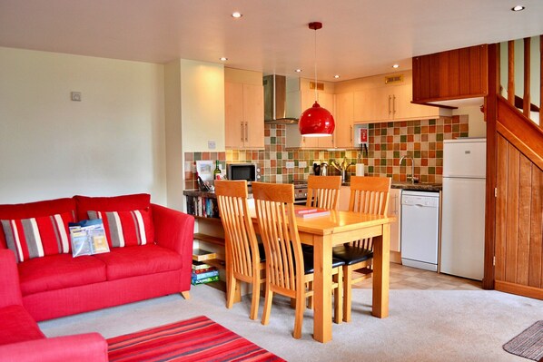 A compact living space with a red sofa, wooden dining table, and chairs, a kitchen with a microwave, toaster, and dishwasher, and a staircase leading to an upper level. - Cottage in Devon Near Dartmoor National Park (Newton Abbot)