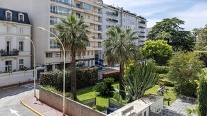 Un immeuble résidentiel à plusieurs étages, doté de balcons, d’un jardin bien entretenu avec des palmiers et d’un ciel dégagé.