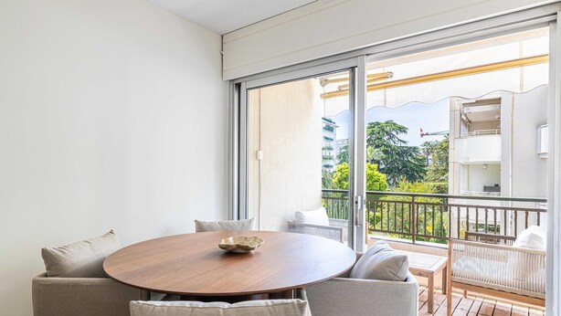 Un balcon avec une table et des chaises en bois, un canapé en osier et une vue sur un immeuble et la verdure.