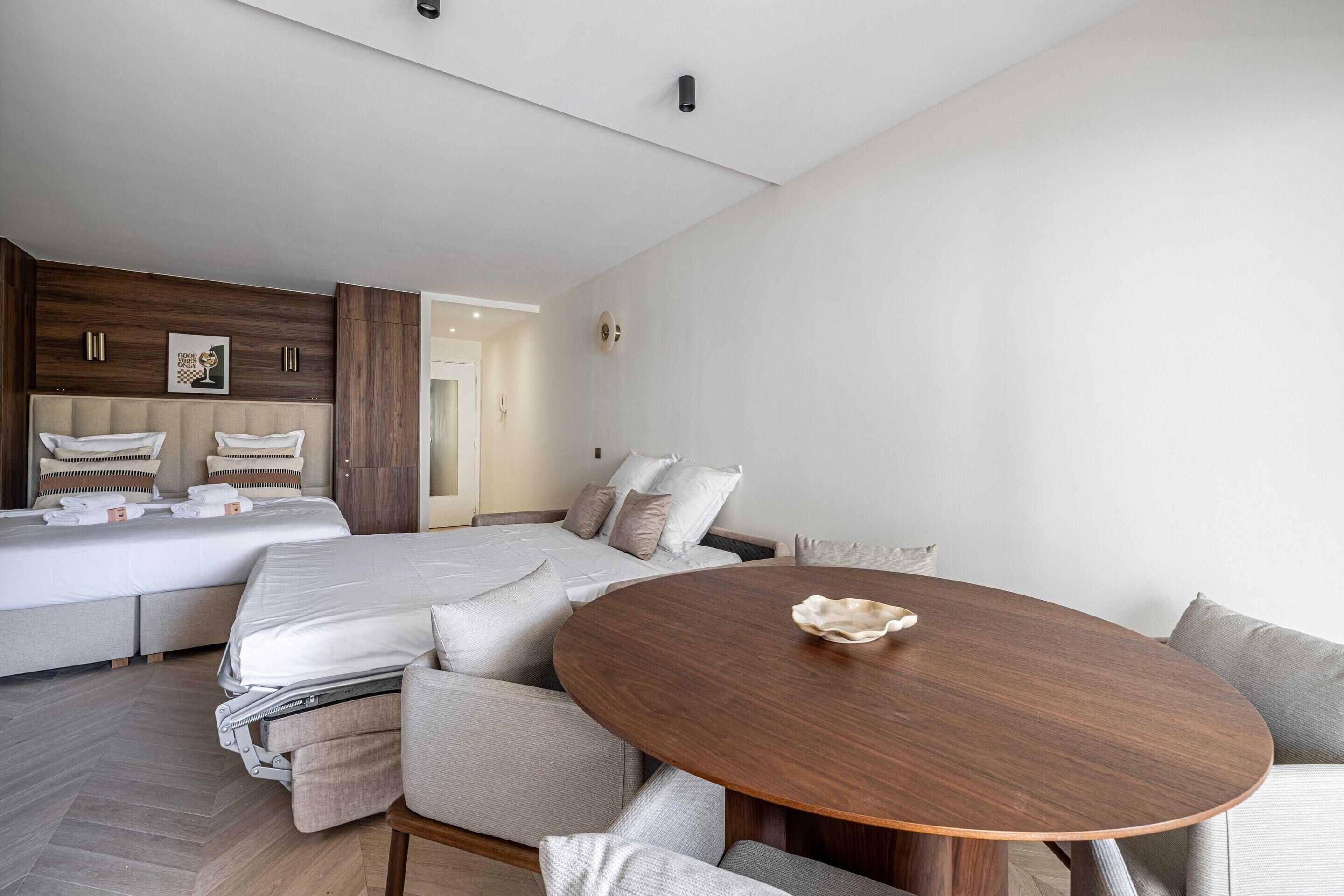 Una habitación de hotel moderna con una cama grande, un sofá, una mesa redonda de madera y una pequeña zona de cocina.