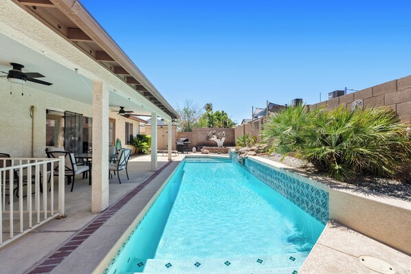 Una piscina rectangular con borde de azulejos, rodeada por un patio cubierto con sillas y mesas.