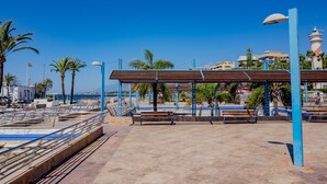 Paseo marítimo con bancos, una zona de estar cubierta y vistas al mar y edificios al fondo.