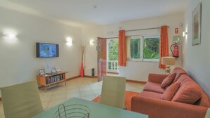 A living area with a sofa, a table, a TV, a bookshelf, and a window with curtains. - Monte Laranja Apt in Lagos (Lagos)
