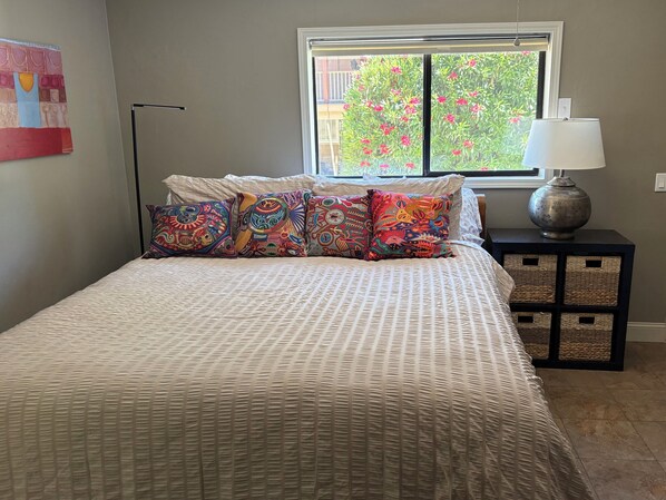 Un dormitorio con cama, almohadas coloridas, ventana, lámpara y una obra de arte en la pared.