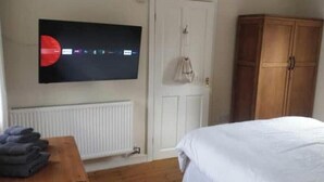 Un dormitorio con una cama, un armario de madera, un radiador, un televisor montado en la pared y una mesa con ropa doblada.