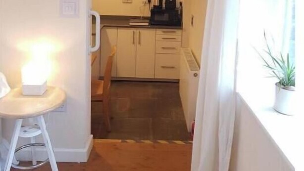 Una cocina compacta con armarios blancos, un pequeño comedor con una silla blanca y una ventana con cortina.