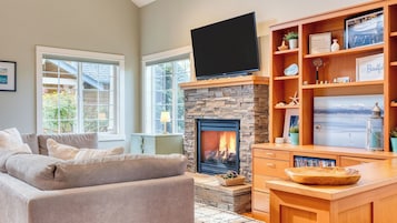 A living room with a stone fireplace, a flat-screen TV mounted above it, a wooden coffee table, and a bookshelf with various items.