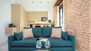 A teal sofa with pink cushions, a coffee machine, and two wine glasses on a white table. A kitchen with a TV and cabinets is visible in the background.