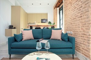 A teal sofa with two wine glasses and a magazine, a kitchen in the background, and a brick wall.
