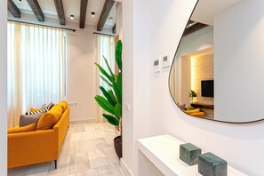 Un salon moderne avec un canapé jaune, une grande plante en pot et une console blanche ornée d’objets qui imitent le marbre.