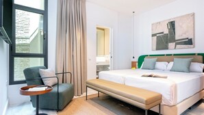 Ein modernes Hotelzimmer mit einem großen Bett, einer Bank, einem Sessel, einem Nachttisch, einem Fenster mit Vorhängen, einem Bild an der Wand und Blick ins Freie.