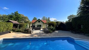 Una piscina con bordo piastrellato, circondata da un giardino e un edificio con tetto rosso.