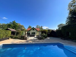 Una piscina con borde de azulejos, rodeada por un jardín y un edificio de techo rojo.