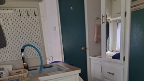 A compact kitchen with a gas stove, a sink, and a small countertop. There is a door with a handle and a mirror above it. - Cute RV in the  forested mountains at Cedar Ridge,  Sonora for your vacation. (Sonora)