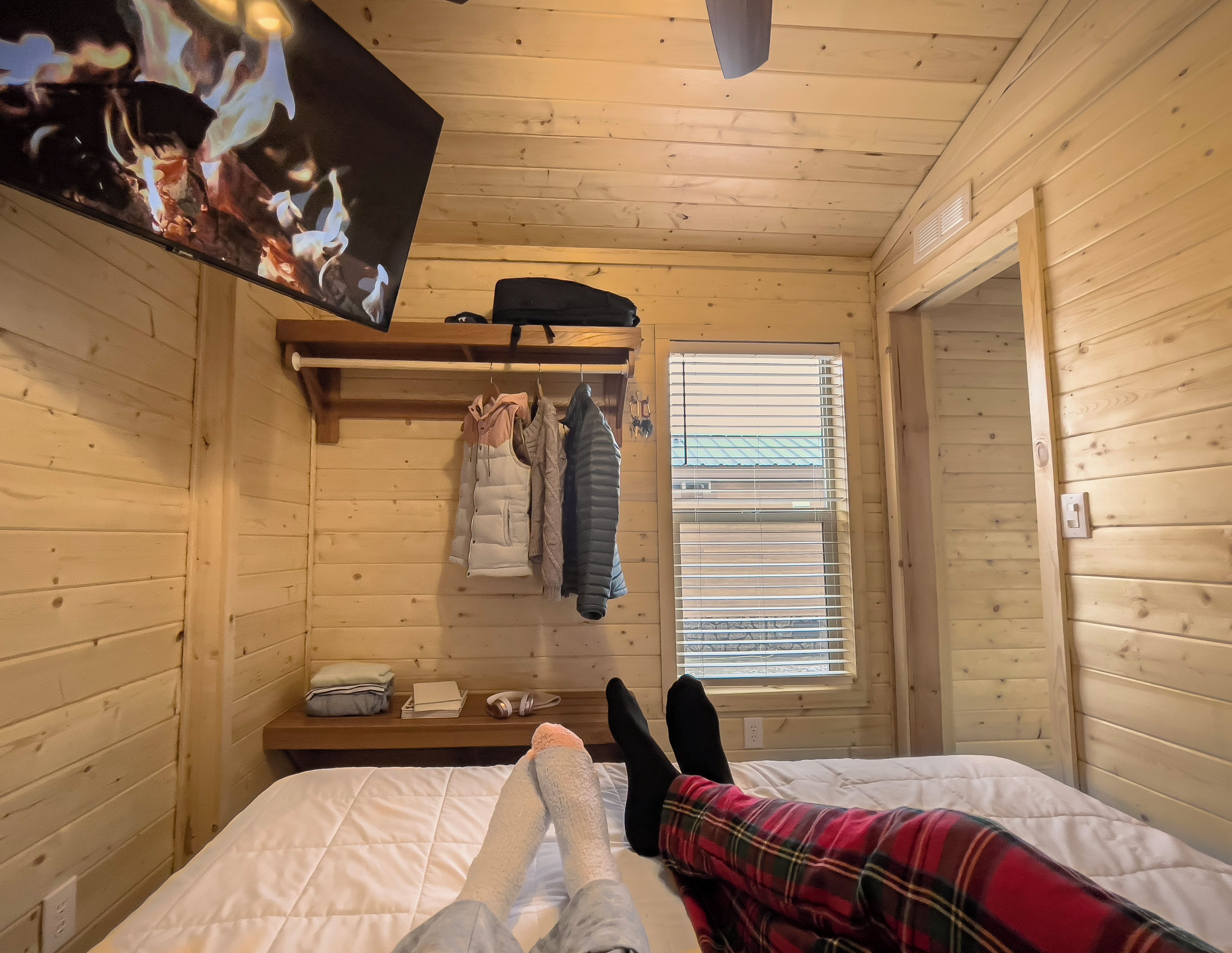 Une pièce de style cabane en bois, avec un lit, une étagère avec des vêtements, une fenêtre avec des stores et un téléviseur fixé au mur.