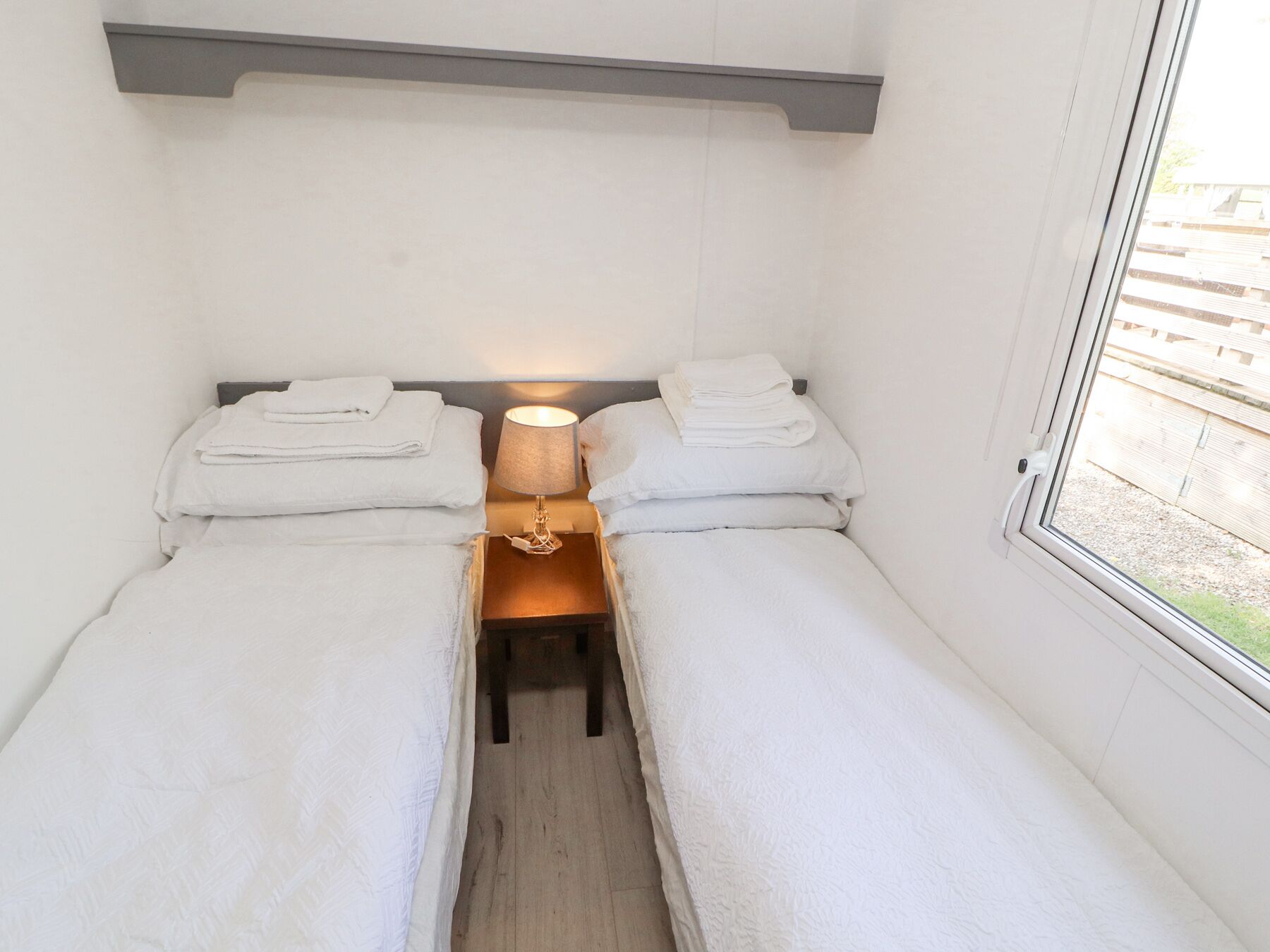 A small, clean room with two single beds, a bedside table with a lamp, and a window with a view of a wooden deck outside.