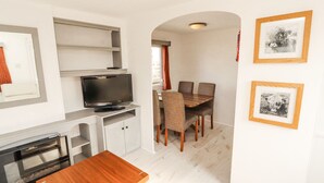 Une cuisine compacte dotée d’un four encastré, un coin repas avec une table et des chaises en bois, et un téléviseur posé sur une étagère.