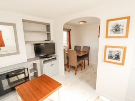 A compact kitchen with a built-in oven, a dining area with a wooden table and chairs, and a television on a shelf.