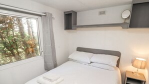 Een net opgemaakt bed met witte lakens en handdoeken, een nachtkastje met een lamp, en een raam met uitzicht op bomen.