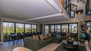 A modern living room with a sectional sofa, a round coffee table, and a dining area with chairs and a table. Large windows provide a view of the outdoors. - Forest Lodge (Pontyclun)