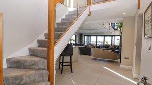 A spacious living area with a staircase, a seating area with sofas and chairs, and a view of the outdoors through large windows. - Forest Lodge (Pontyclun)