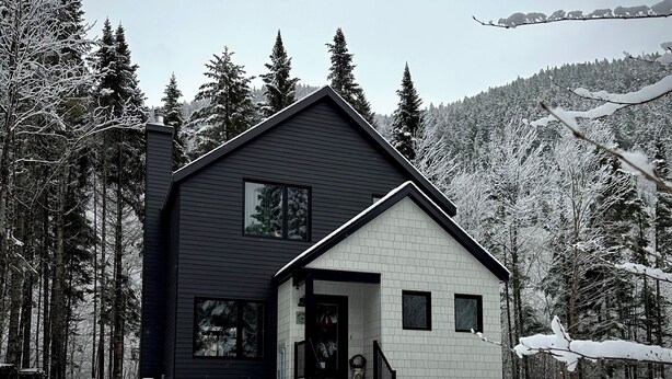 A modern two-story house with a dark exterior and white lower section, surrounded by snow-covered trees and a forested hillside. - Modern chalet 2 minutes from the Bras-du-Nord Valley | Spa & Sauna  (Saint-Raymond)