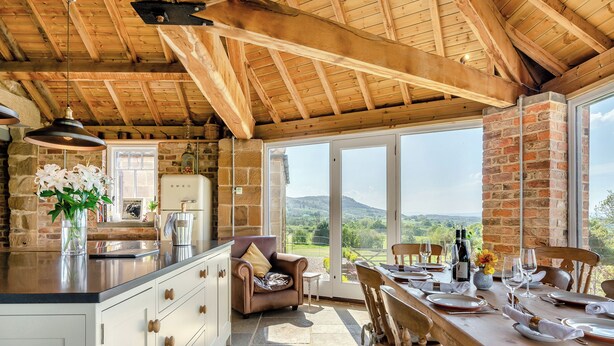 Una cocina rústica con encimera de piedra, vigas de madera y vistas al campo a través de amplias ventanas.
