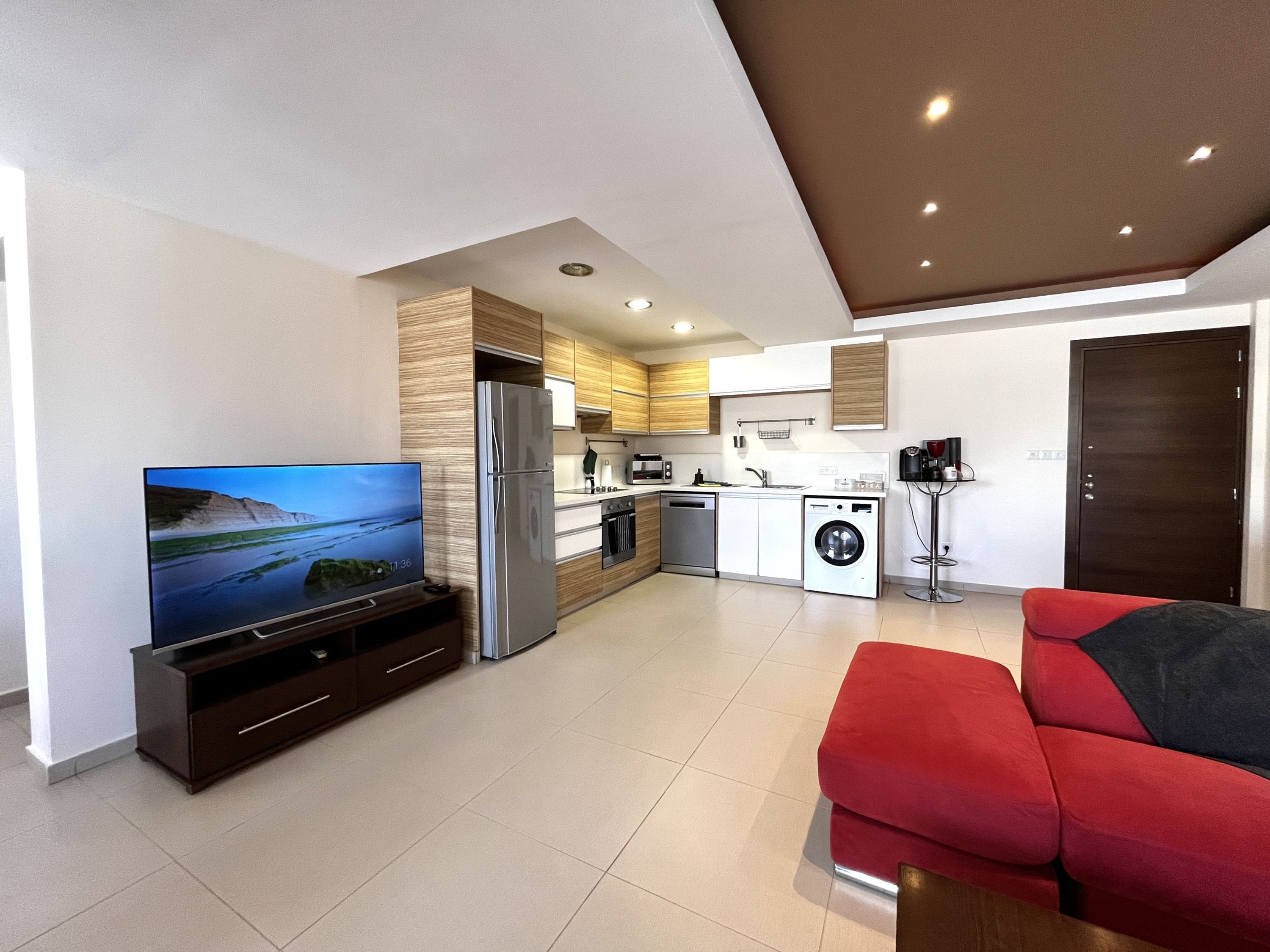 Una cocina moderna con horno empotrado, refrigerador y lavadora. Hay un televisor de pantalla plana sobre un mueble bajo y un sofá rojo en la sala.