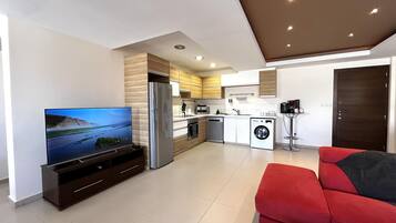 Una cocina moderna con horno empotrado, refrigerador y lavadora. Hay un televisor de pantalla plana sobre un mueble bajo y un sofá rojo en la sala.