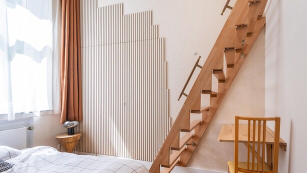 A bedroom with a wooden staircase, a bed with a white and black geometric-patterned bedspread, a wooden nightstand, and a chair. - Apartment in Koblenz Near Deutsches Eck (Koblenz)