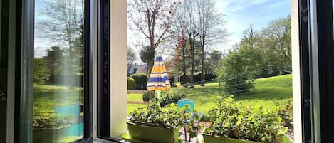 Ventana con vista a un jardín, que incluye césped, árboles y un paraguas colorido.