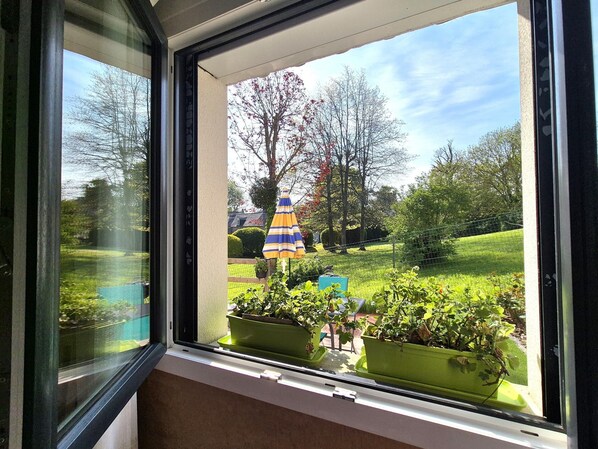 Une fenêtre donnant sur un jardin, avec une pelouse, des arbres et un parasol aux couleurs vives.