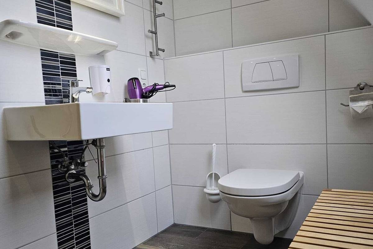 A modern bathroom with a white sink, mirror, and toilet. There is a wooden bench and a towel rack.