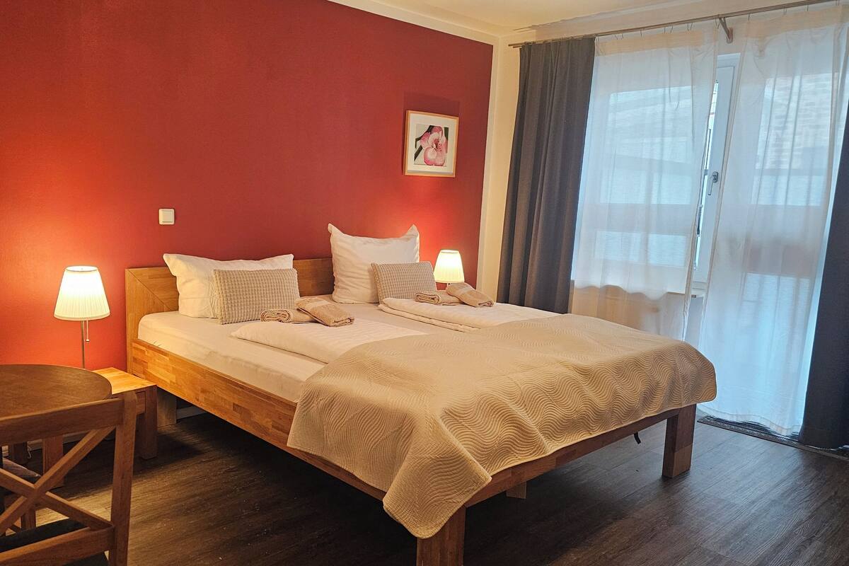 A hotel room with a wooden bed, two bedside lamps, a wooden chair, a small table, a red wall, and a window with curtains.