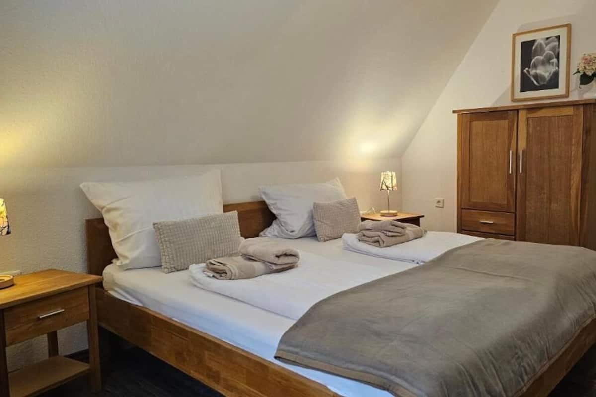 A neatly made bed with pillows and a blanket, a wooden nightstand with a lamp, and a wooden wardrobe with a framed picture.