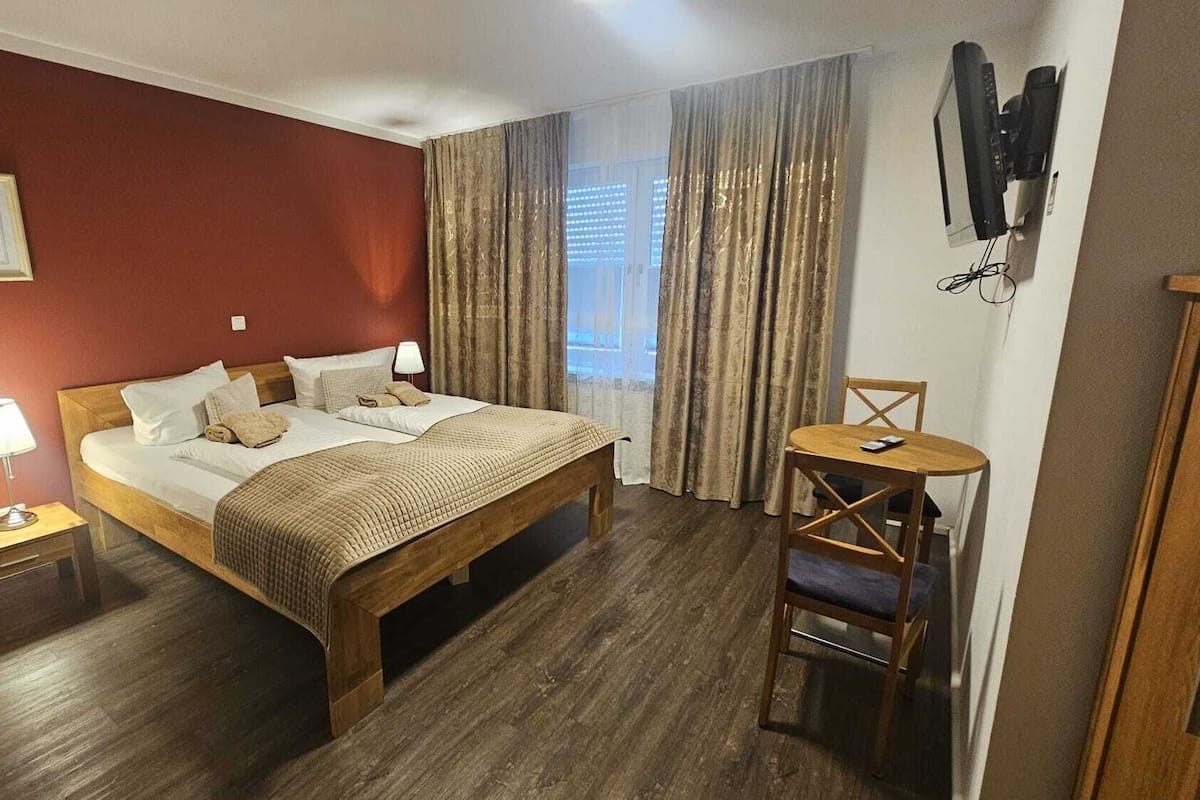 A hotel room with a wooden bed, a bedside table with a lamp, a chair, a TV mounted on the wall, and a window with curtains.