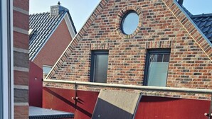 A view through a window of a room with a wooden deck, a red wall, and a brick building with a circular window. - Mondlicht Norddeich (Norden)