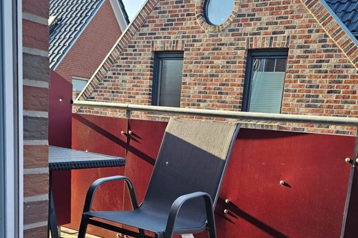 A view through a window of a room with a wooden deck, a red wall, and a brick building with a circular window.