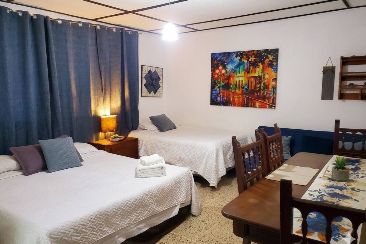 A hotel room with two beds, a dining table, a lamp, a painting, and a small plant.