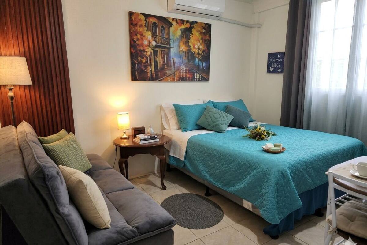 A bedroom with a bed, a sofa, a nightstand, a lamp, a painting, and a window with curtains.