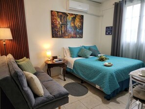 Un dormitorio con cama, sofá, mesita de noche, lámpara, un cuadro y una ventana con cortinas.